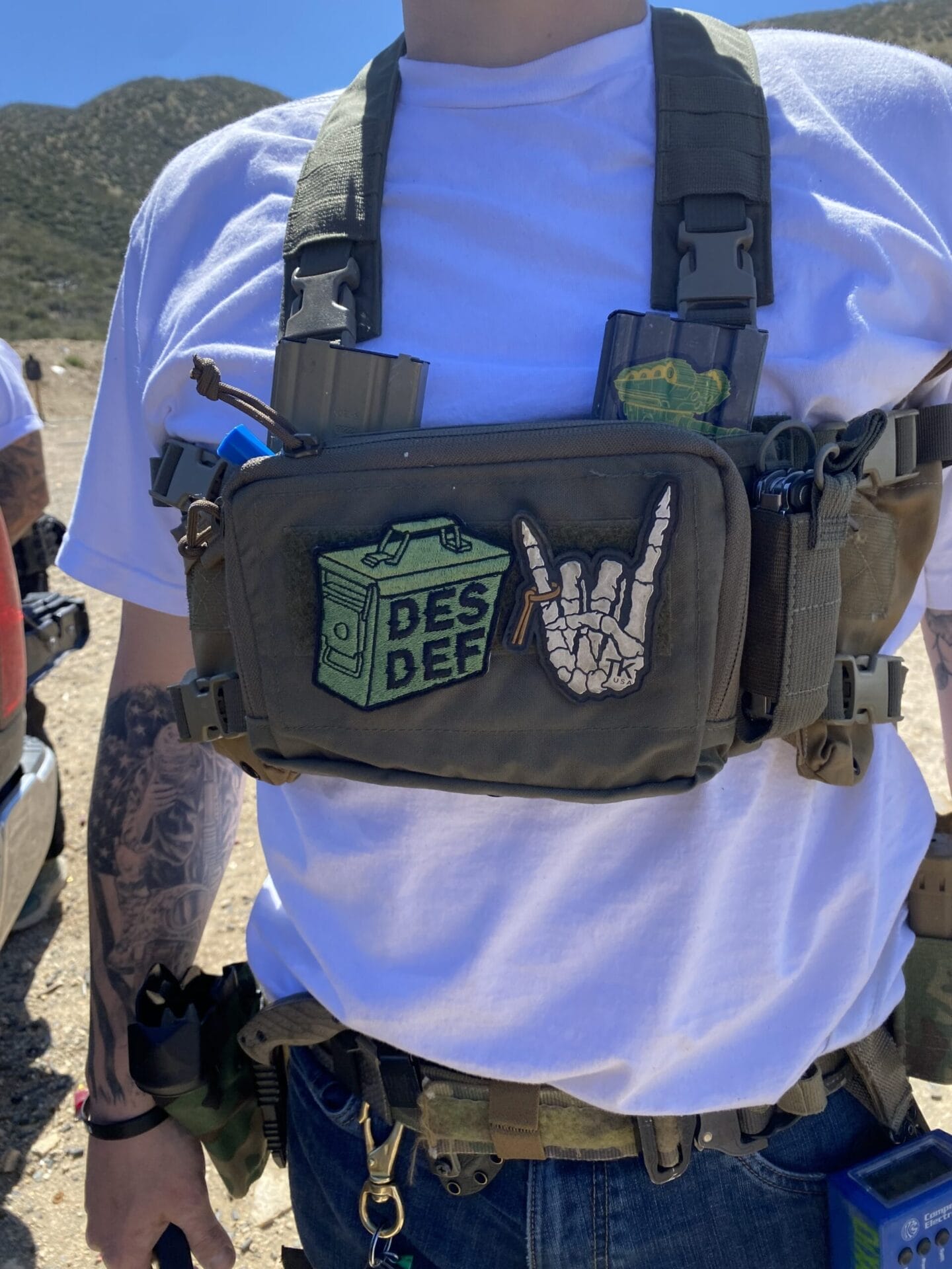 Person wearing a white t-shirt and tactical vest with patches, including one that reads "DES DEF" and another with a rock hand gesture, standing outdoors in a mountainous area.